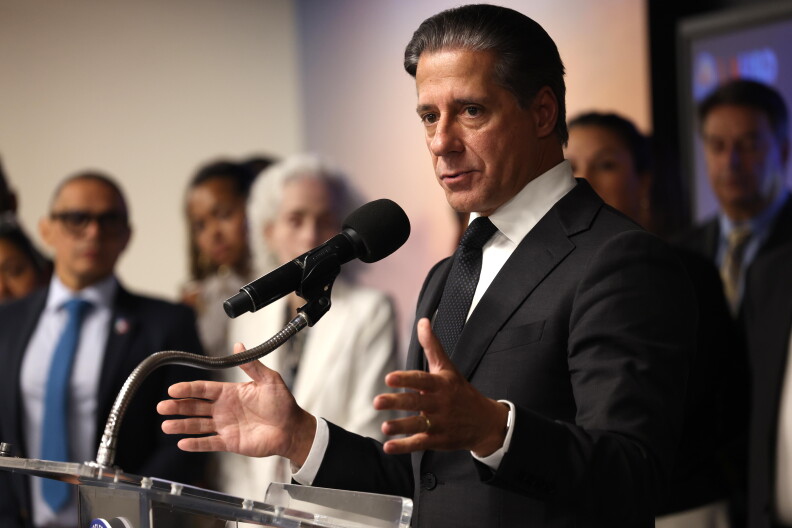 A man with medium light skin tone wears a dark suit and tie and speaks into a microphone at a podium. A number of adults in business clothes can be seen behind him in the background.