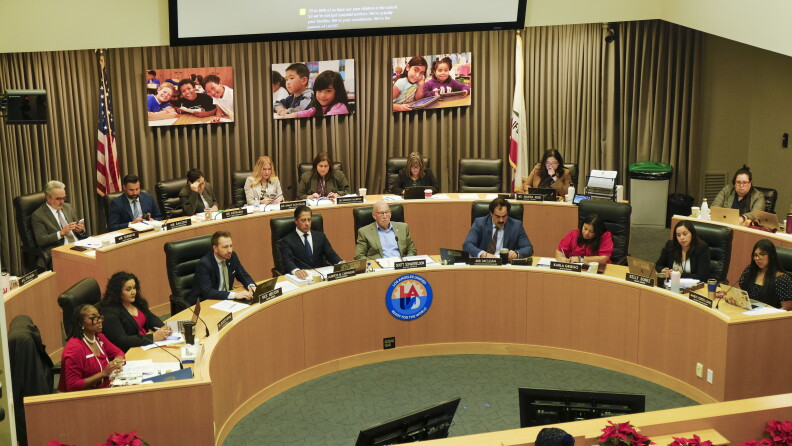 Nine people sit at a curved light brown wood dais. From left to right there is a woman with dark skin tone, dark brown hair and a red jacket, a woman with medium light skin tone and dark brown curly hair, a man with light skin tone, light brown hair and a beard, a man with medium skin tone wearing a navy blue suit with a tie and white shirt, a man with light skin tone, white hair, and glasses in an olive green sport coat, a man with dark brown hair, a mustache and a blue sport coat with a brown tie, a woman with medium light skin tone, dark brown hair and a red dress, a woman with medium light skin tone and a black blazer and a teenage girl with a dark brown long hair and a black polka dot shirt on. There is a logo on the dais that reads LA.
