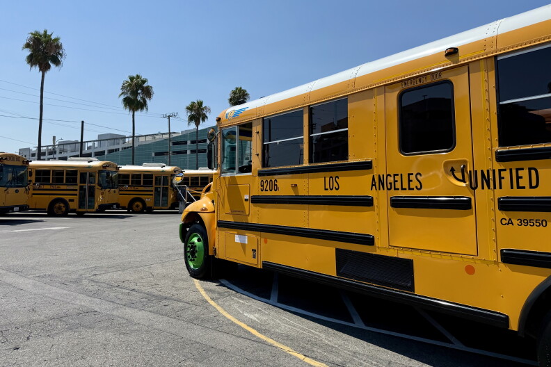 A yellow school bus with green wheels is a parked next to several other buses. The side of the bus reads Los Angeles Unified and there are palm trees in the background. 