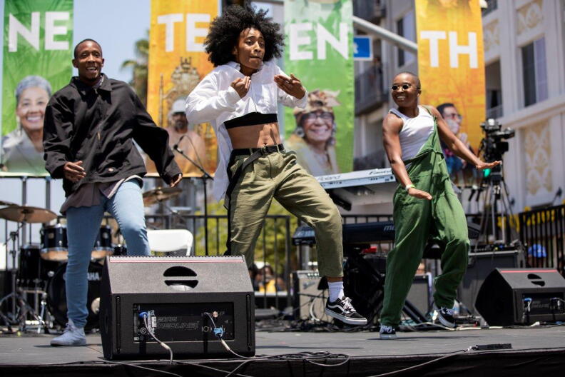 Three people dancing on a stage at an outdoor event. Behind them are colorful banners with partially visible text, along with musical instruments like drums and keyboards. The stage includes speakers and cables in the foreground.
