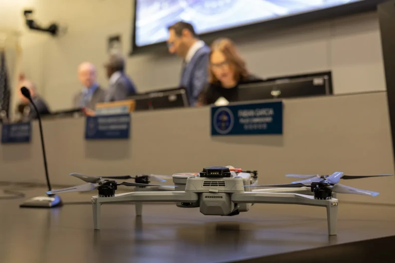 A small drone is set on a table in the foreground in front of a row of nameplates and people talking amongst themselves out of focus in the background.