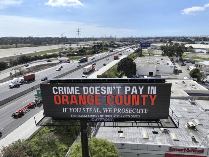 A billboard with the words "crime doesn't pay in Orange County" in white and orange letters. The billboard stands along a freeway with multiple cars and big rigs on it.