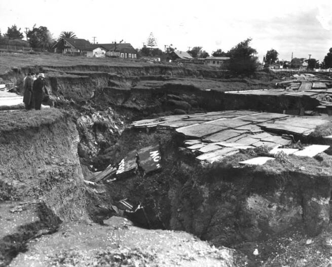 A black and white archival look at a large section of ground that's cracked off from the bluff. The debris clearly shows paved roads as two people stand on the edge looking over. They are much smaller compared to the size of the crack.