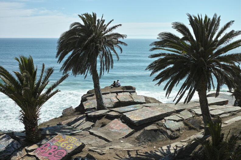 A wide look at a cliffside with the blue Pacific ocean in the background. The cliff is filled with broken concrete, griffiti and palm trees. Two people are sitting on the edge.