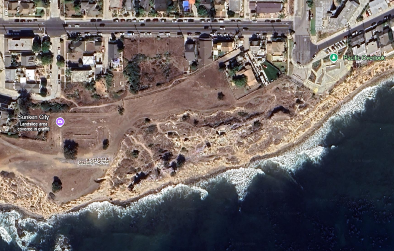 A satellite aerial view of the landslide area. The cliffside is visible from above, with a clear breakage on the edge. The inland area is flat and unbroken, and further in are homes and the neighborhood.