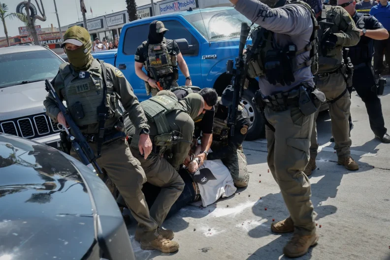 Federal agents with face coverings arrest a man on the floor as more agents and a police officer stand around them in between vehicles.