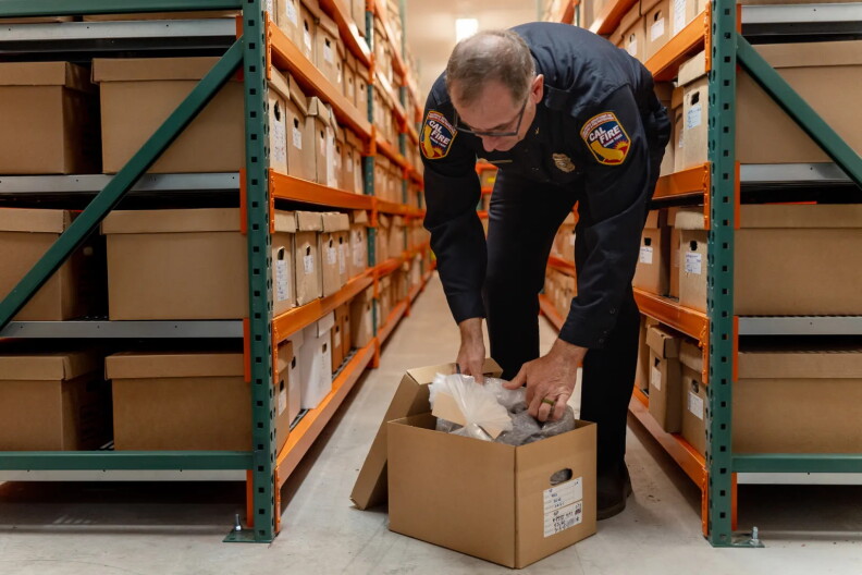 A uniformed Cal Fire employee examines materials inside an open archival storage box in a records warehouse. Long rows of metal shelving filled with labeled cardboard boxes line both sides of the aisle, creating a corridor-like view through the archive.