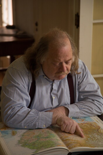 A male presenting person sits at a table. He points to an open book that shows a map.