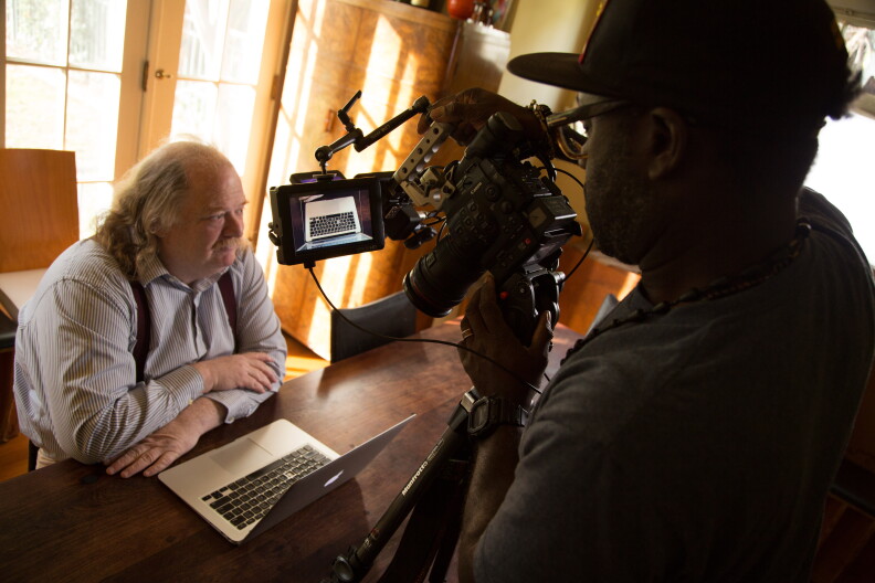 A male presenting person sits at a table with a laptop open in front of him. A film camera is set up in front of him.