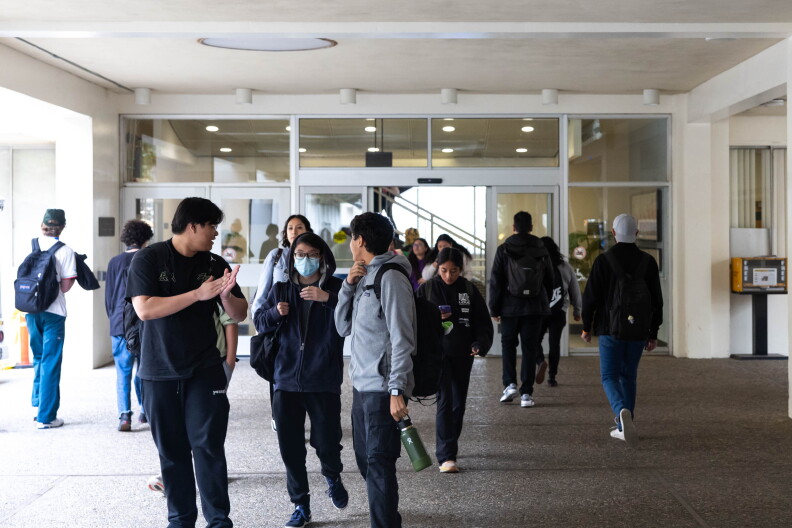 A group of diverse students wearing backpacks walk out of a white building with glass doors.