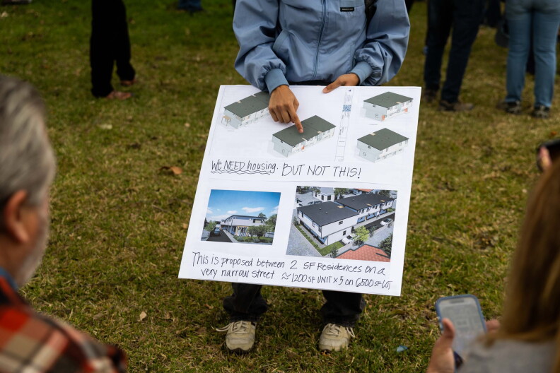 A person holds a poster and points to a rendering of a multi-unit building. The sign reads "We need housing, but not this!"