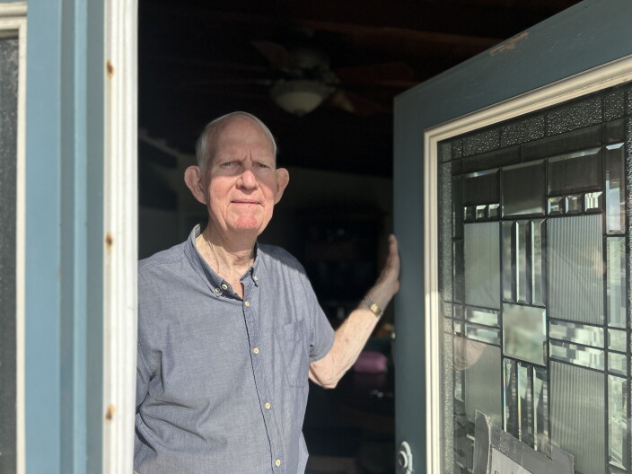 An older man with light skin tone wears a gray-blue collared buttonup shirt and stands in his doorway as the sun hits his face. 