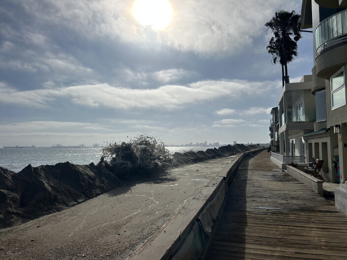 A boardwalk lined with apartments facing sand berms and a wave overtopping them. The sky is blue and white clouds. 