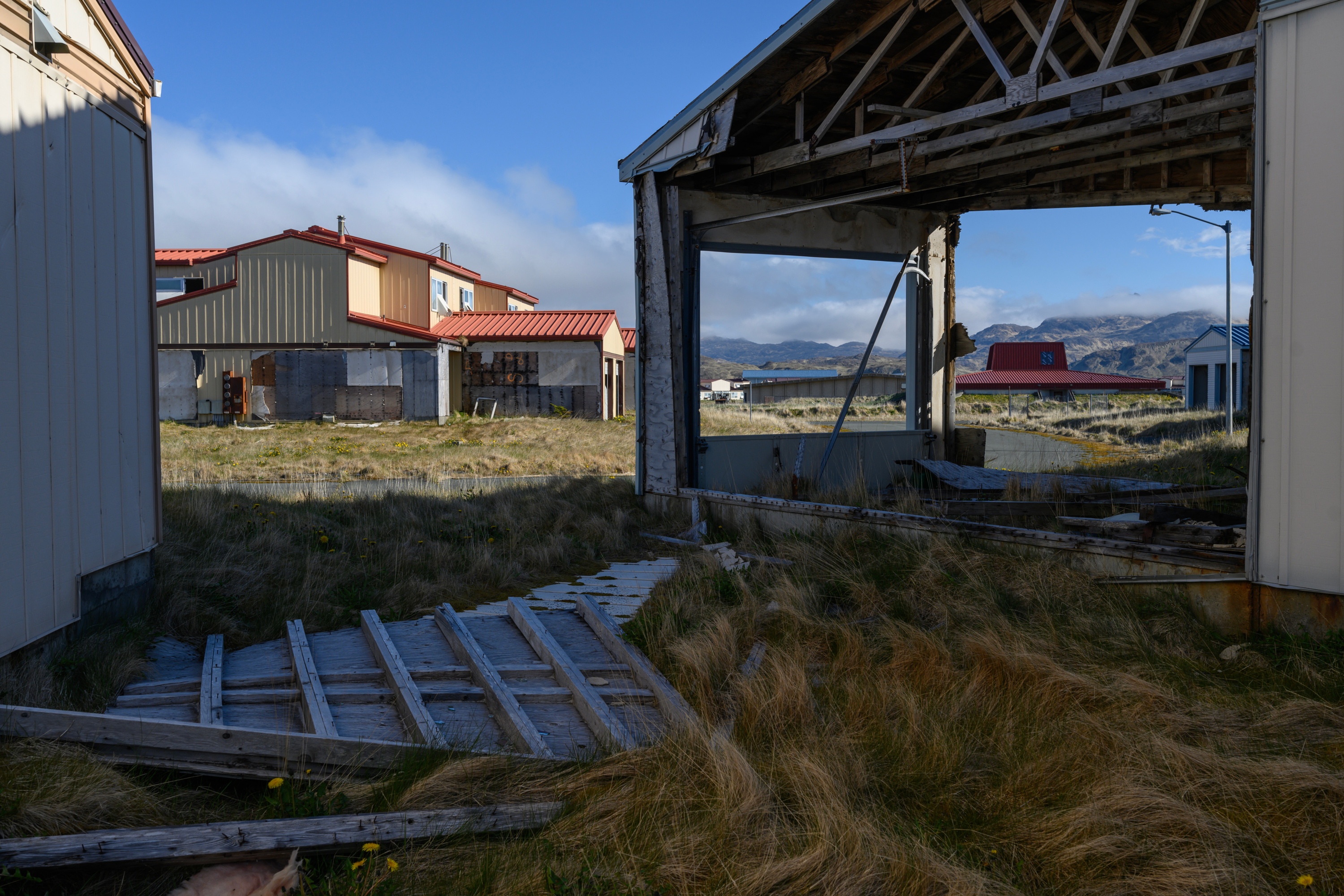 Brown, overgrown grass covers a fallen wall on the ground, and the viewer can see straight through the frame of a house in the foreground. In the background, a two-story house has missing wall panels. The sky is blue, and white clouds and mountains can be seen behind the houses.