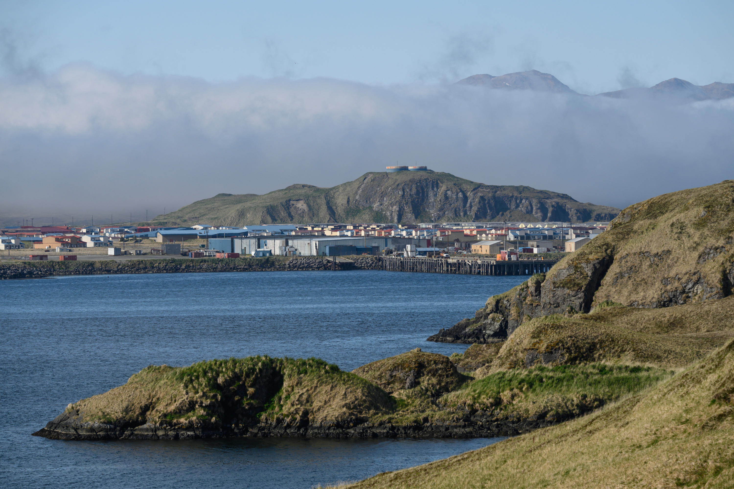 Gray clouds hang over a green island with small houses and buildings, surrounded by blue water.