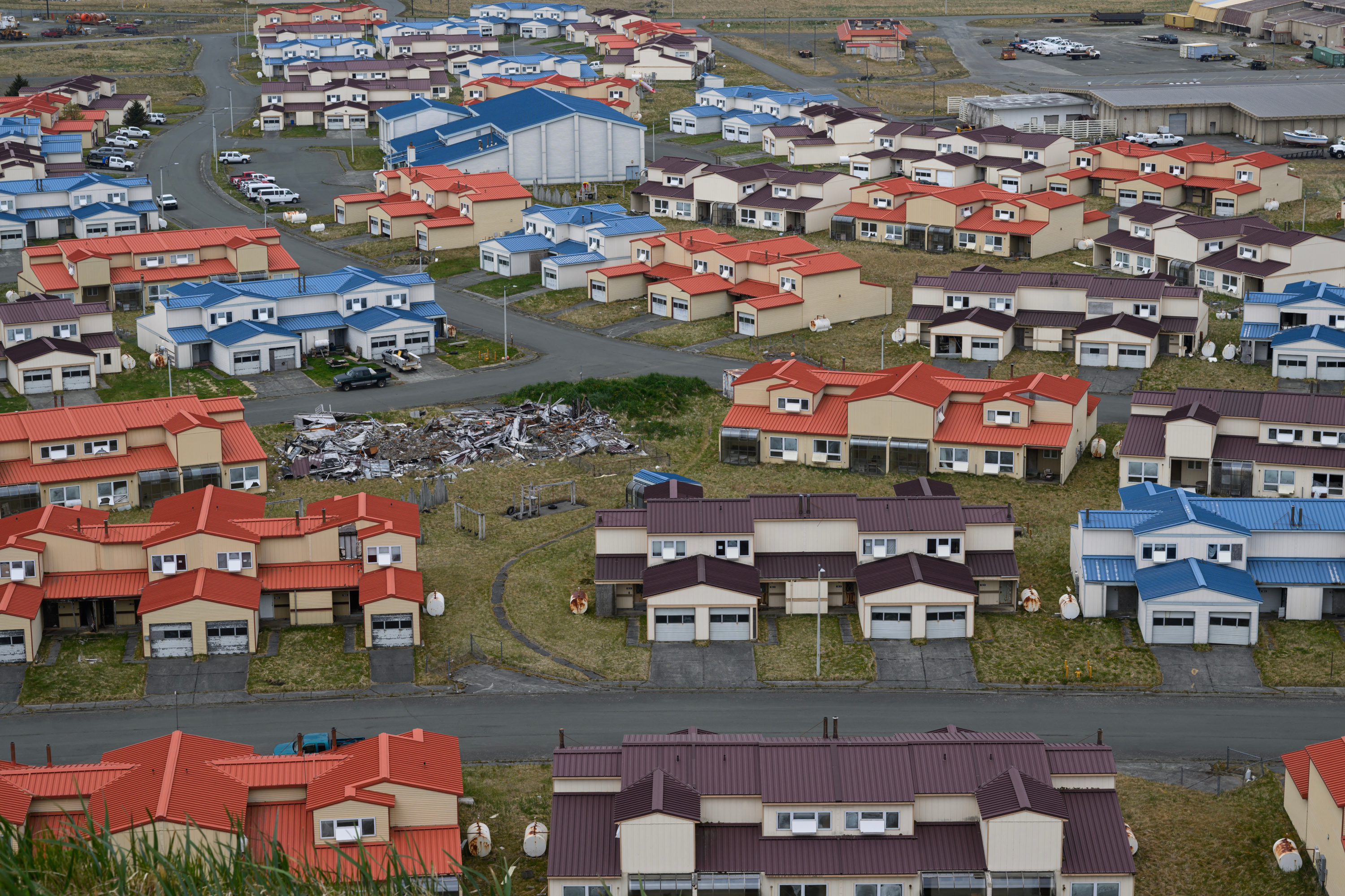 Aerial view of a subdivision of houses with brown, red and blue roofs. One lot is covered in debris. A few cars are parked throughout the neighborhood.