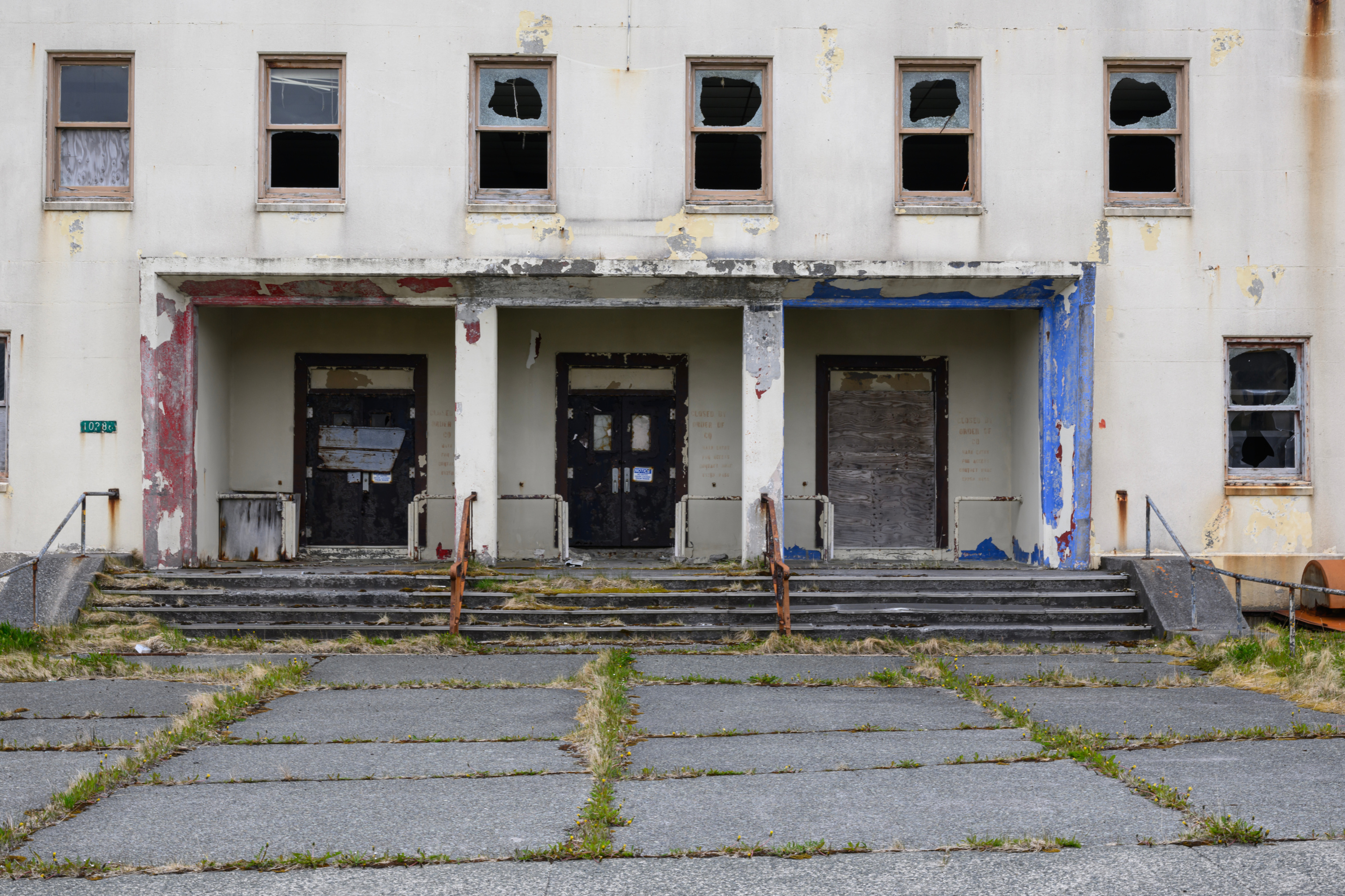Stairs covered in weeds lead up to a beige two-story building with shattered and boarded-up windows, boarded-up doorways and peeling paint.