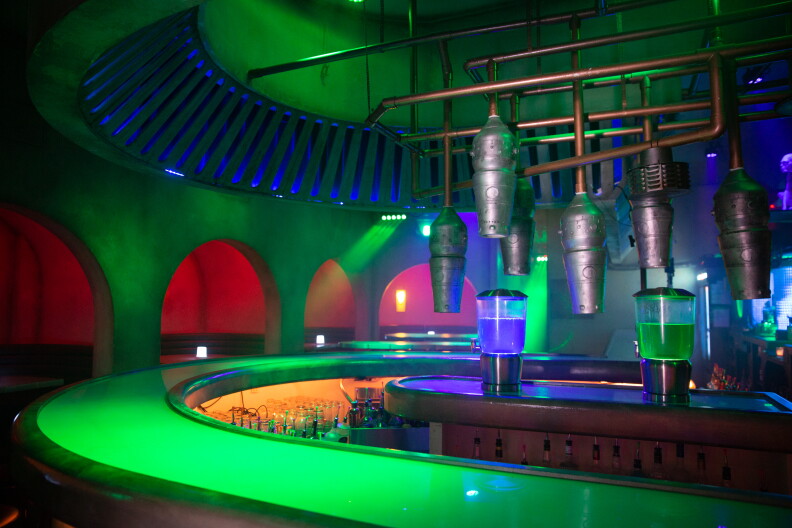 A spookily lit interior with a circular bar. The bar has green lights, the ceiling green and blue and areas behind arches are red and orange.