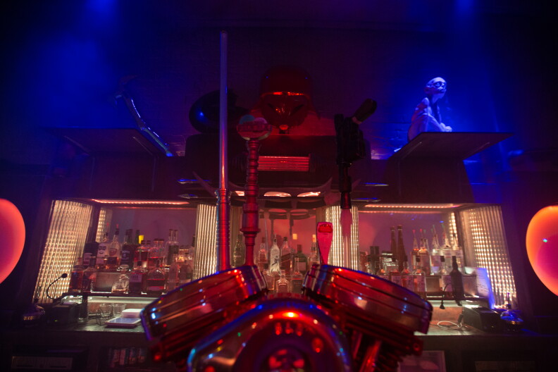 A spooky looking interior of a bar; in the foreground is a red metallic phone; in the background behind the bar are rows of bottles, and above alien creatures are lit in blue.
