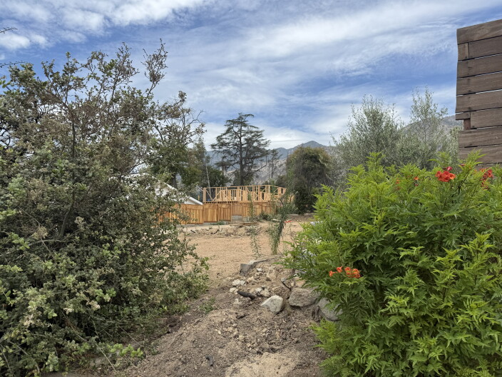 Wood framing of new construction rises on a dirt lot with overgrown bushes. Trees and mountains are visible in the distance under a sky with patchy white clouds.