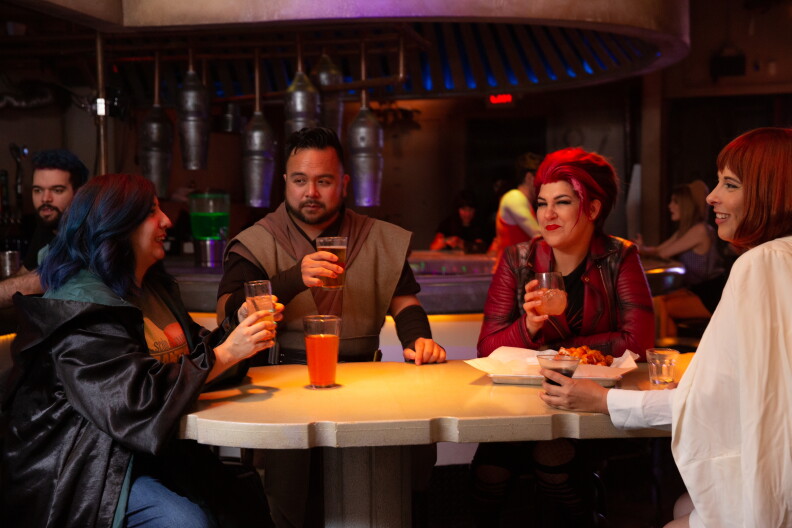 Three light skinned women and a medium skinned man are sitting around a table. All are wearing different costumes connected to franchises like Star Wars.