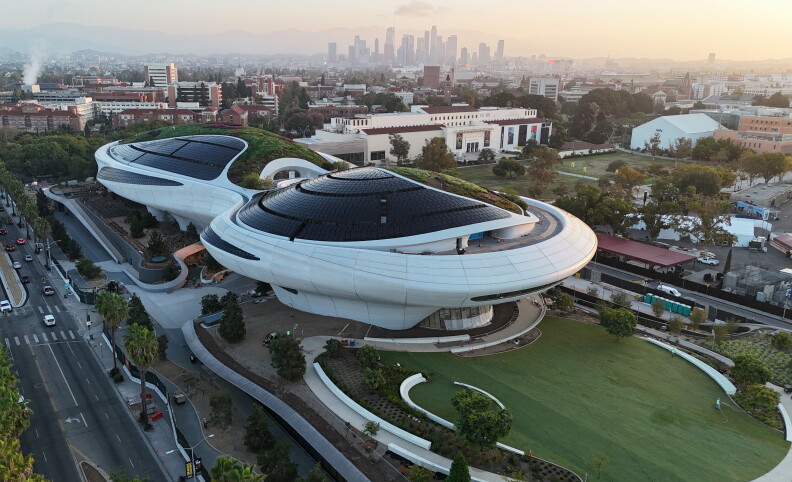 A white building in an infinity shape with black, glass roofing. Off to the left is a street with a few cars driving by. In front the white building is a large grass area.
