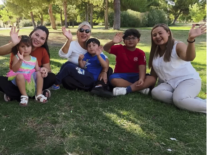 A family with young children wave and smile while sat on grass as part of a news report on Spain's approach to immigration in October 2025
