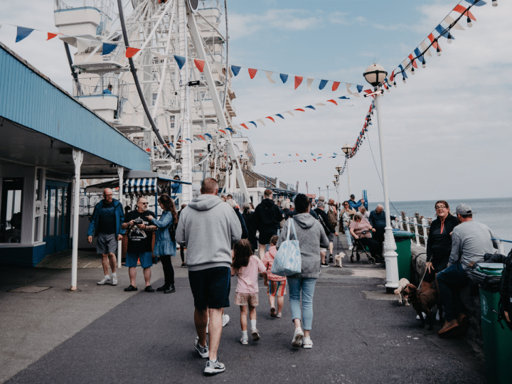 A bustling scene at Llandudno promenade in Wales with people walking near a Ferris wheel adorned with flags. Days out like this will not be affordable for many UK families due to money worries.