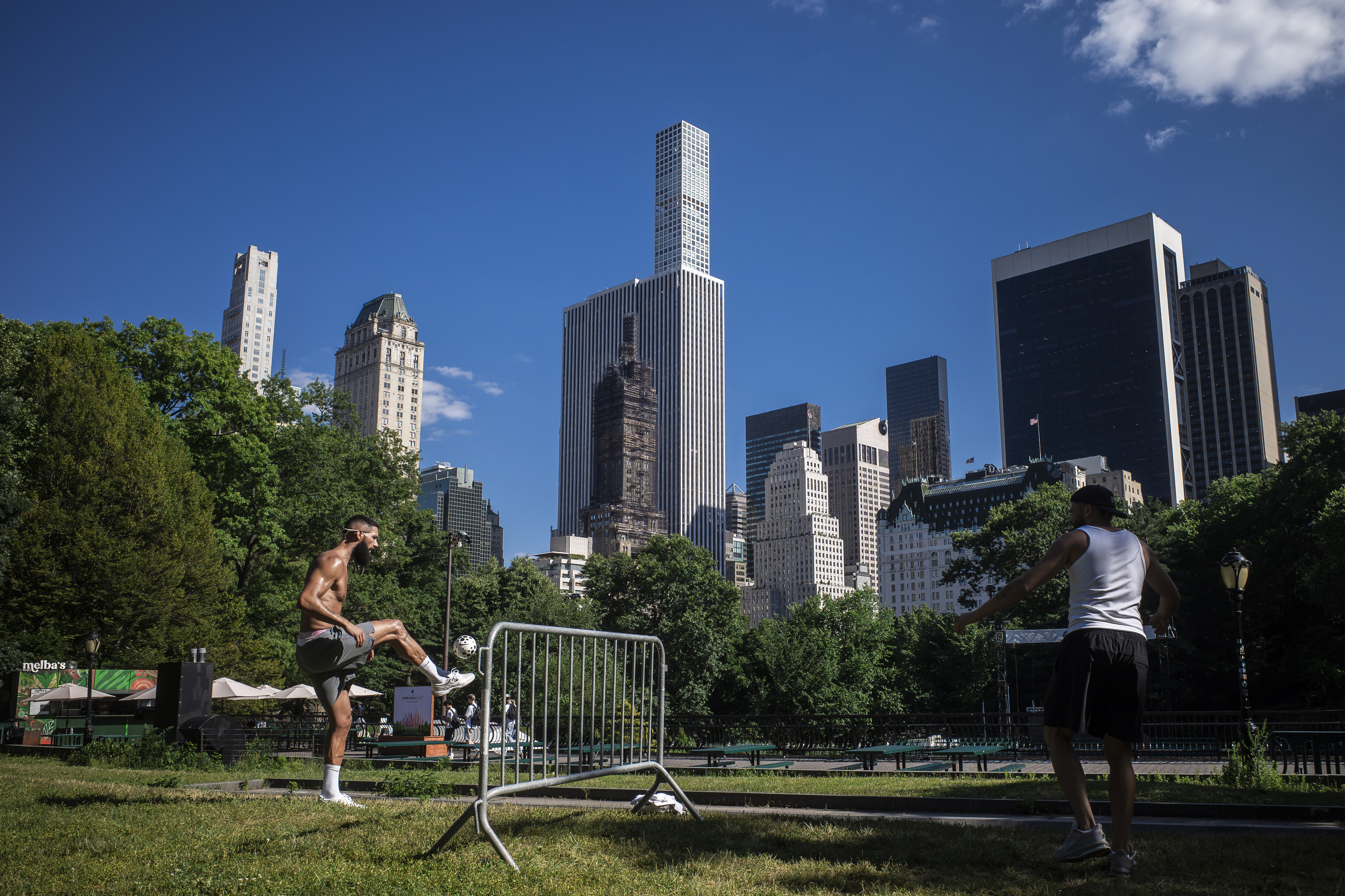 Two men play street football and juggle a ball over a barricade in Central Park on a sunny afternoon.