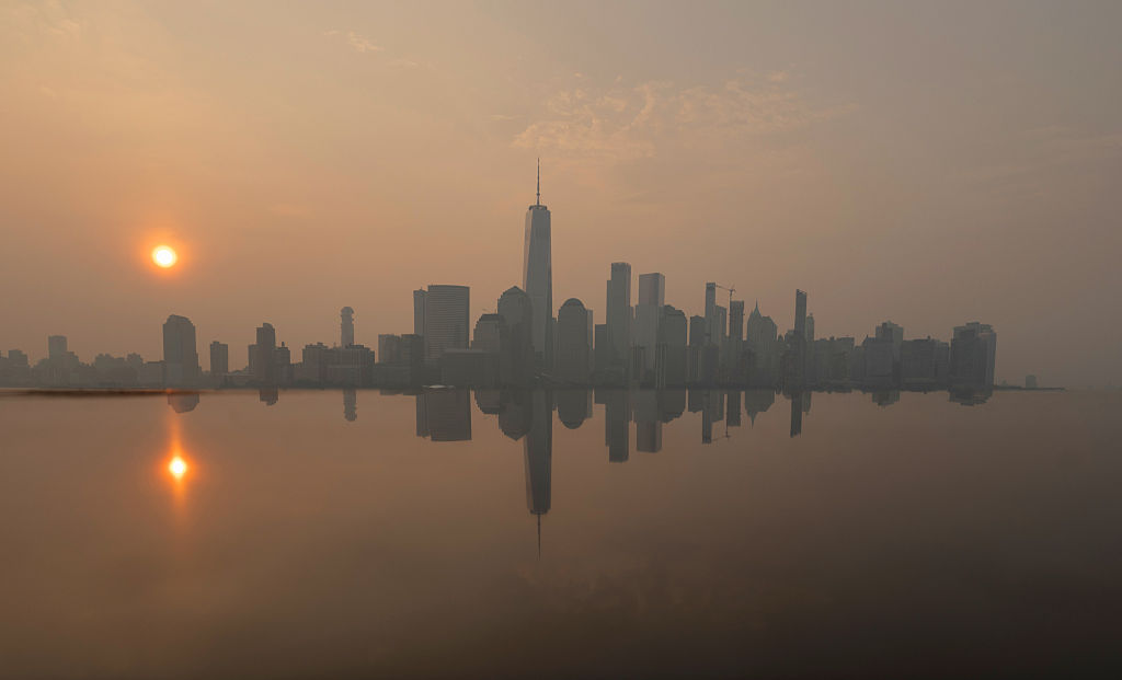 The skyline of lower Manhattan and One World Trade Center are reflected on the top of a monument as wildfire smoke from Canada shrouds the sun as it rises in New York City on July 18, 2026, as seen from Jersey City, New Jersey.