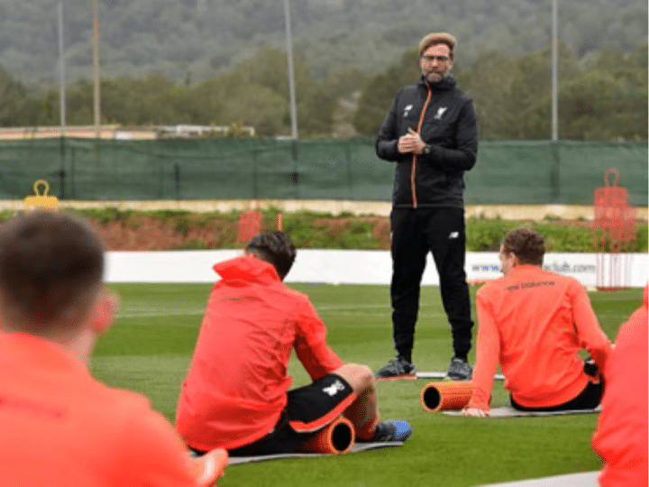 Jürgen Klopp stands to address his football players, who are sat with their backs to camera all wearing the same red jacket, during what he describes as a ‘pre-season training’ camp in La Manga in February 2017