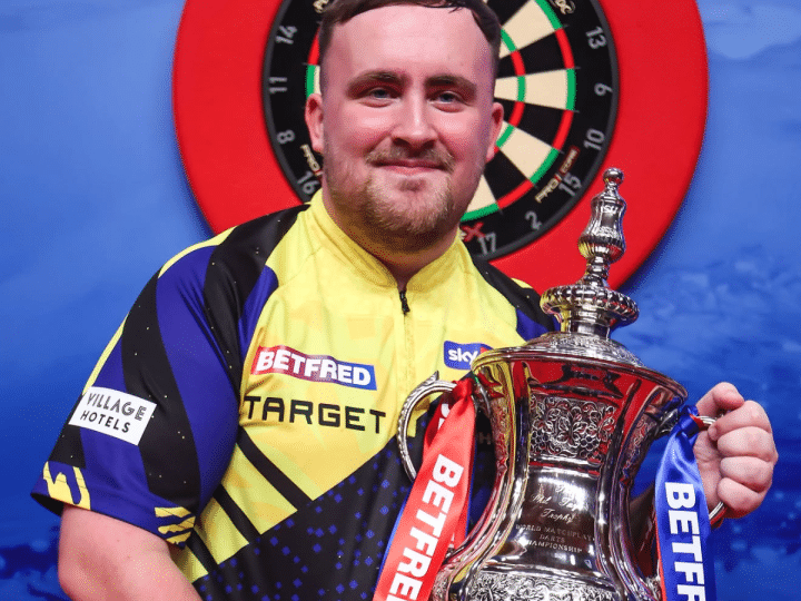 Luke "the Nuke" Littler smiles with his winner's trophy in hand infront of a darts board in Blackpool