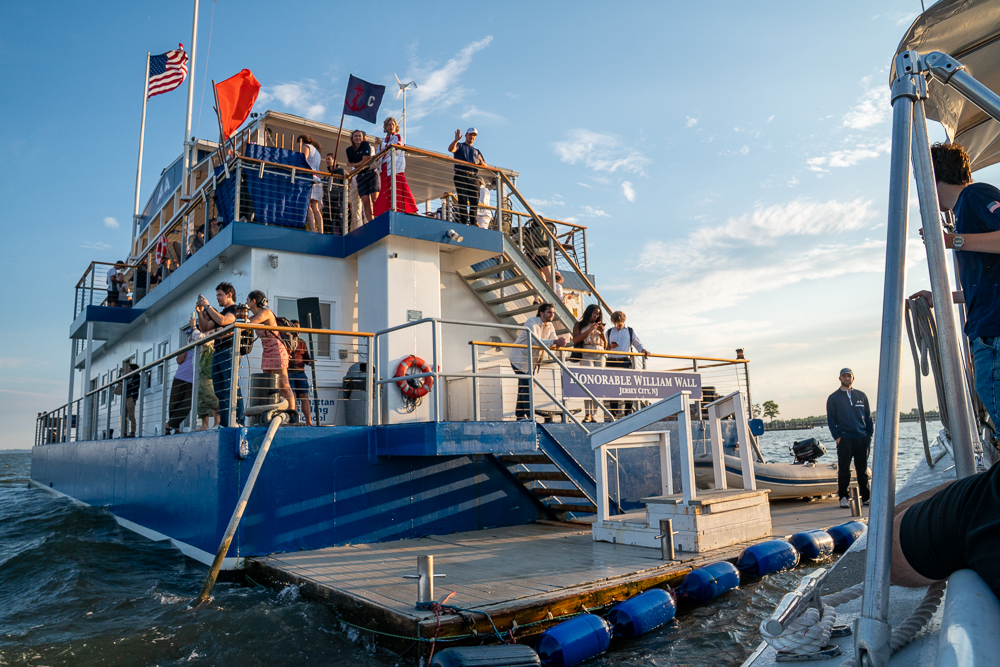 The exterior of a bar on a barge in a river with people waving and flags flying.