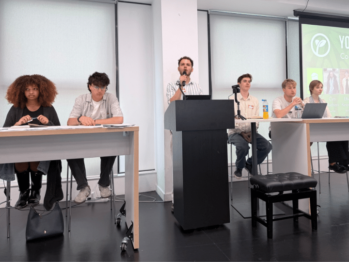 Young Greens party conference. Each candidate is sat at the front of a classroom on two desks with a podium and mic in the middle, where one candidate is speaking