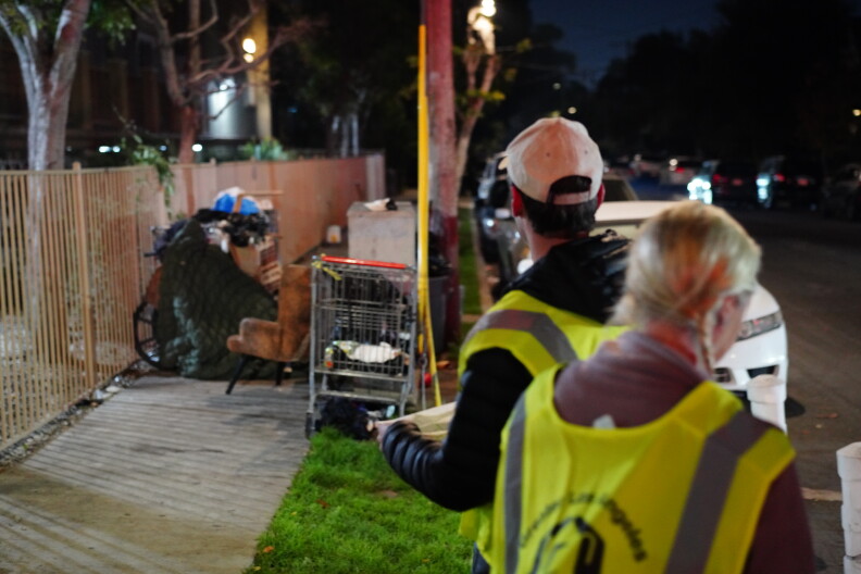 Two people wearing reflective vests stand next to a makeshift shelter on the sidewalk.