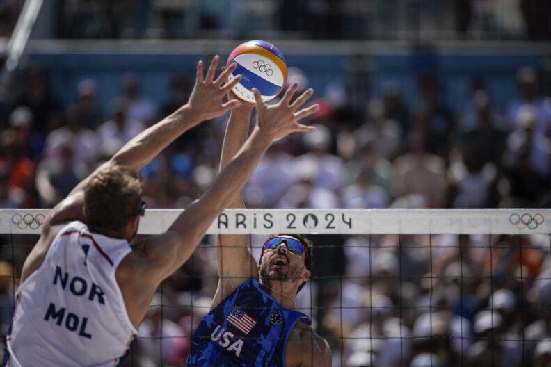 Two volleyball players jump right in front of the net, one spiking the ball and the other trying to block it.