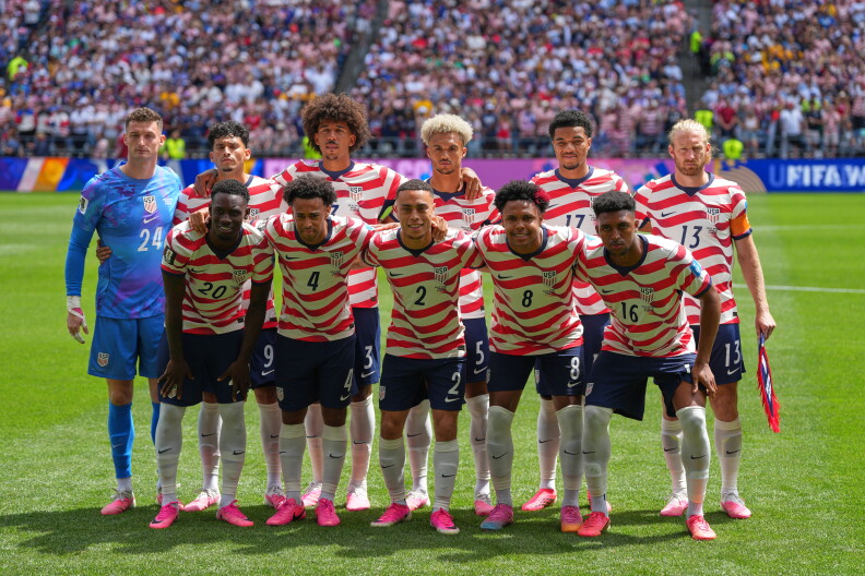 Two rows of men mostly in red and white striped uniforms have their arms around each other on a soccer field.