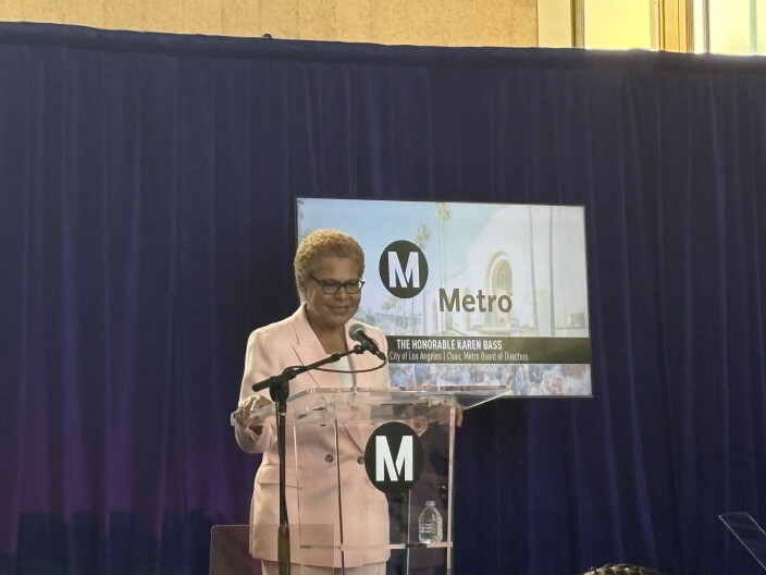 A woman with short brown hair stands in front of a podium with a microphone. The woman is wearing a light pink suit and a white shirt underneath. The podium is emblazoned with the letter "M" in white in a black circle. Behind the woman is a screen that has the same "M" logo and "Metro" written in black. There's a black banner at the bottom of the screen that reads"The Honorable Karen Bass" in all caps.