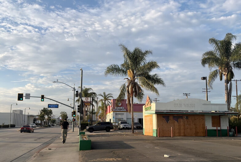 A person on an electric scooter drives past a boarded up white and green building that still has markings indicating that it was once a car wash. Palm trees and a cloudy sky are in the background of a residential area.