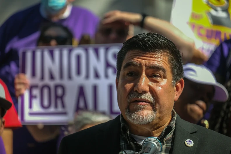 David Huerta, a man with medium skin tone with a gray beard, speaks into a microphone. There are people behind him out of focus holding up signs, with one that reads "Unions for all."
