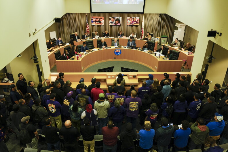 Dozens of people stand before a dais where a group of people sit.