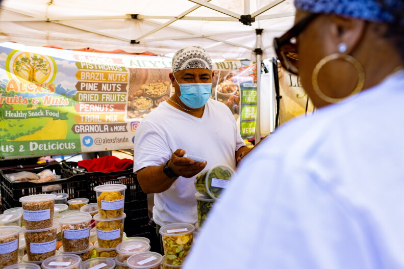 A vendor at the Crenshaw Farmers Market sells a variety of fruits and nuts. He wears a surgical mask, a white t-shirt.