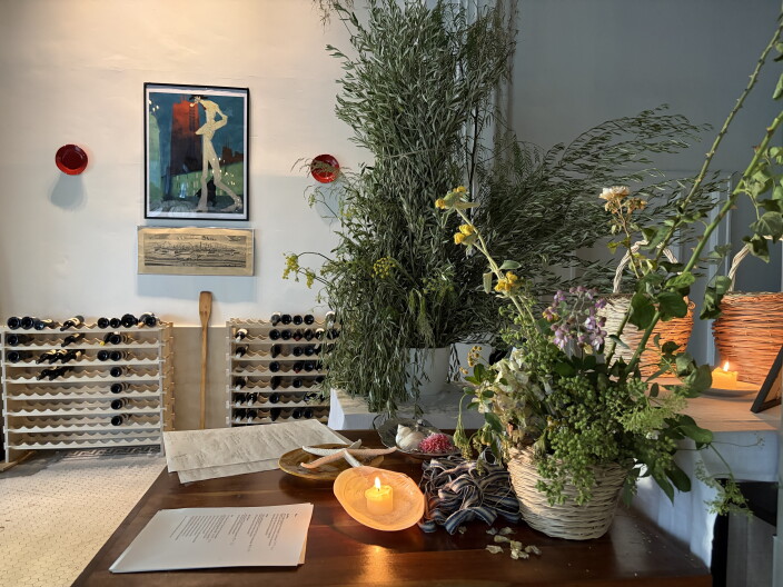 The interior of a restaurant has a hostess desk in the foreground, with a lit candle in an oyster shell and various pots with greenery on it; in the background are wine racks with bottles of wine and a colorful modernist painting on the wall.