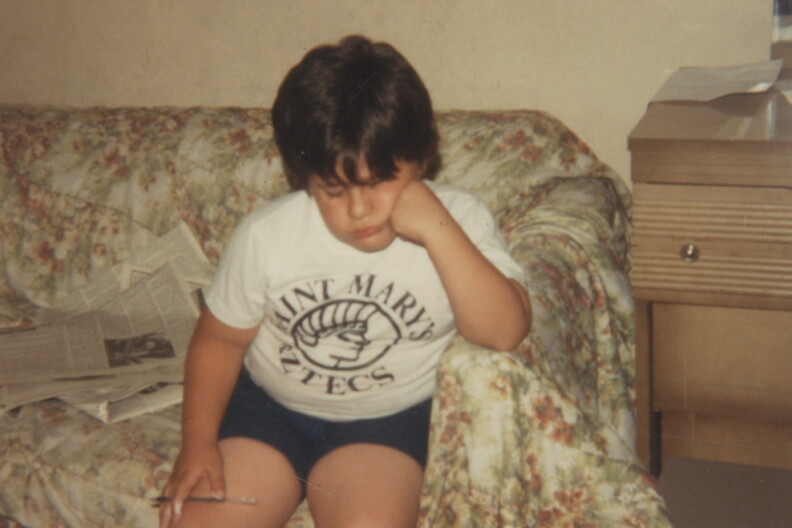 A childhood snapshot a boy with brown hair, resting his chin in his hand as he sits on a floral-print couch holding a pencil. He wears a white "Saint Mary's Aztecs" T-shirt, with newspapers spread out beside him.