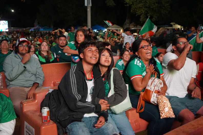 A crowd watches a World Cup match on a large outdoor screen. Many of them wear green jerseys. A young man and woman hold hands, sitting on a bench.