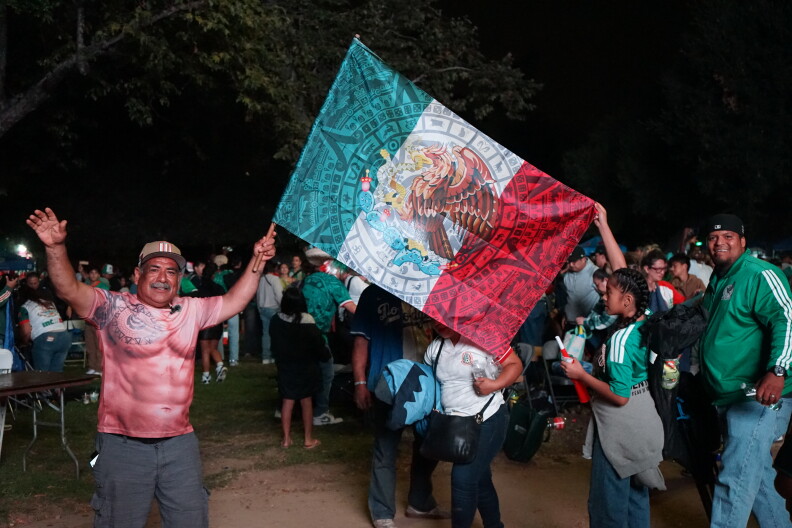 A medium skin toned man holds a large Mexican flag, his arms raised. It's nighttime. A young girl wearing a green Mexico jersey helps him hold the flag.