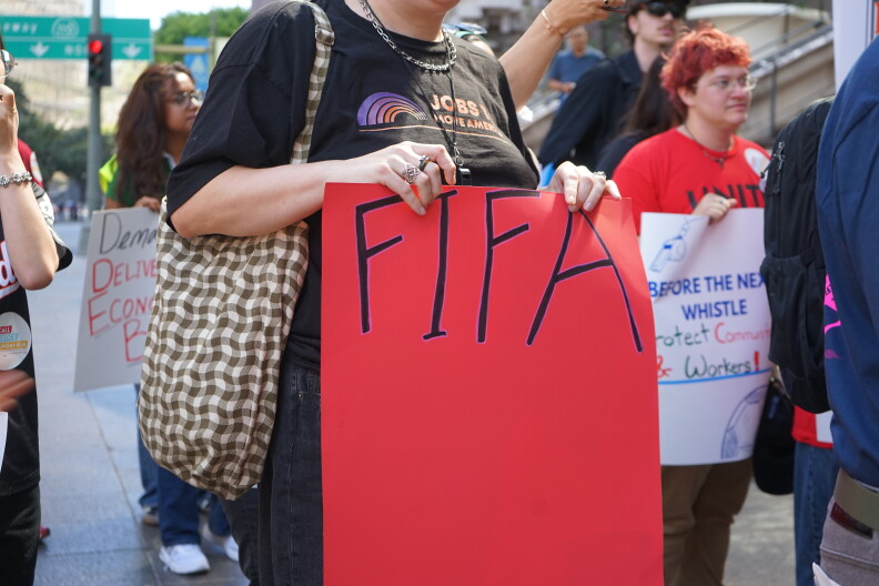 A person with a light skin tone wearing a black t-shirt holds a red poster that reads "FIFA." The image is solely of the person's torso, but behind them you see other demonstrators.