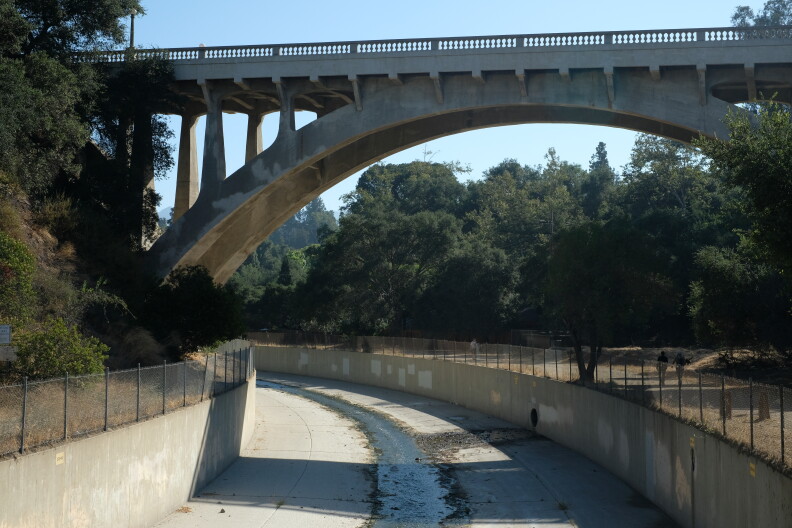 A concrete bridge arches over a channelized stream. Lush trees are on either side.