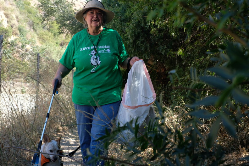 An older woman with light brown skin and a brimmed hat wears a green shirt that reads "Save my home." She carries a plastic trash bag and trash picker in a nature area.