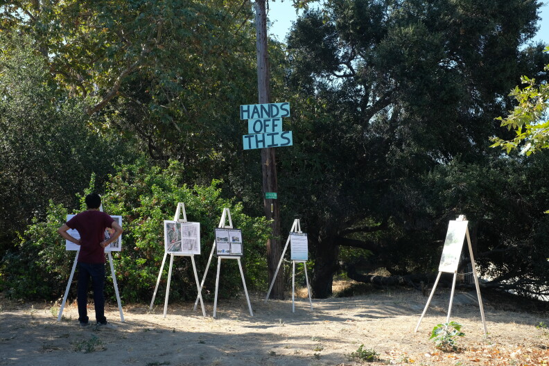 A man faces away from the camera looking at signs propped up around a dirt trail entrance to a wooded area.